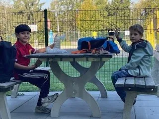 Zwei junge Jungen sitzen an einem Tisch im Park. Ein Junge trägt ein rotes Shirt und der andere ein blaues Jackett. Sie sind von Taschen und anderen Gegenständen umgeben.