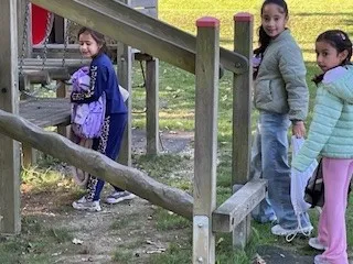 Vier junge Mädchen stehen in einem Outdoor-Spielplatz, eine hält einen Rucksack und eine andere einen Plastikbeutel.