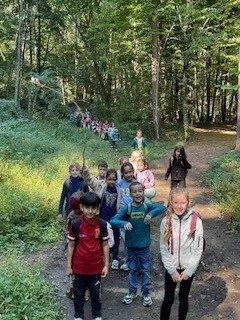 Eine Gruppe von Kindern läuft auf einem Lehmweg im Wald. Einige halten Stöcke und andere haben Rucksäcke.
