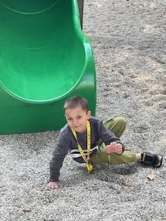 Ein junger Junge in einem grauen Sweatshirt und grünen Hosen sitzt auf dem Boden neben einer grünen Rutsche.