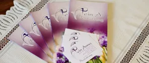 Mehrere gefaltete Broschüren und Visitenkarten mit 'Klein Anna Bestattung' darauf, auf einem weißen Tuch. Die Karten und Broschüren sind lila und weiß.