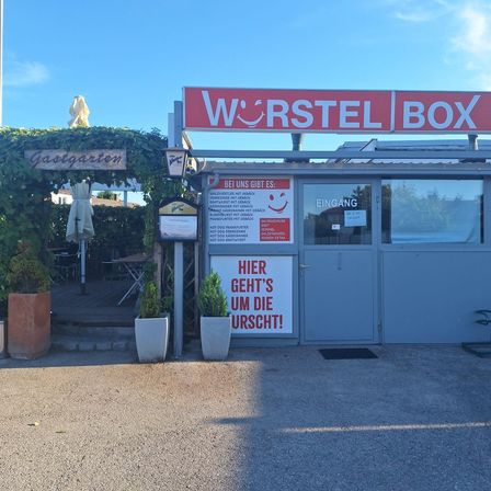 Das Wurstel Box-Etablissement mit einem Eingangsschild, das 'Hier geht's um die Wurst' steht. Topfpflanzen stehen davor.