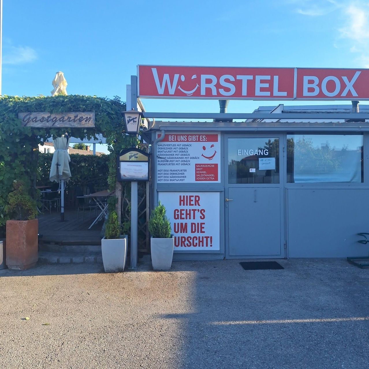 Ein rotes Schild mit weißen Buchstaben sagt Wurstel Box über einer Glastür mit einem Schild, das Hier geht's um die Wurst! sagt. Blumentöpfe und ein geschlossener Schirm sind in der Nähe.