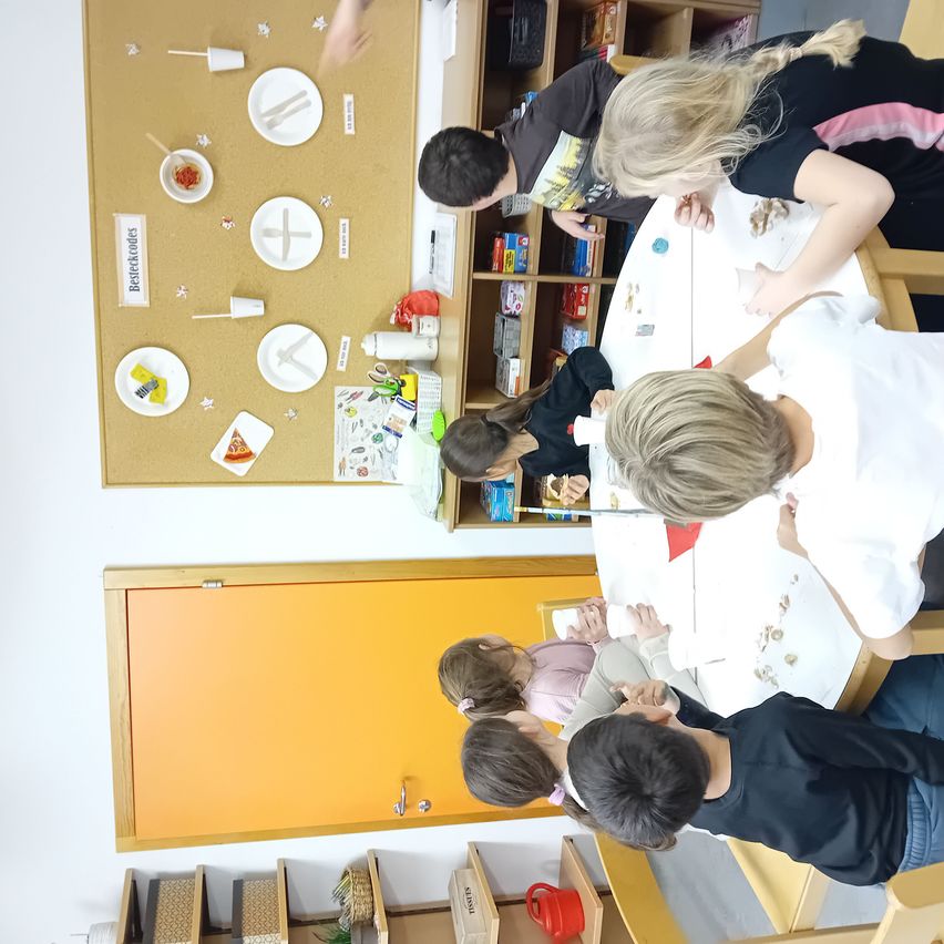 Children are sitting around a table, engaged in an activity. There are plates, cups, and other items on the table. A board with drawings is on the wall.