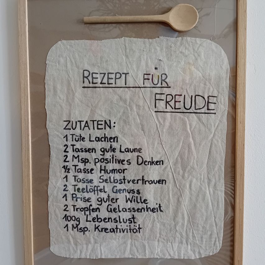 Ein gerahmtes Stück zeigt ein Rezept für Glück, einschließlich Zutaten wie Lachen, gute Gedanken, Humor und Selbstvertrauen. Es ist auf einem Stück Papier geschrieben.