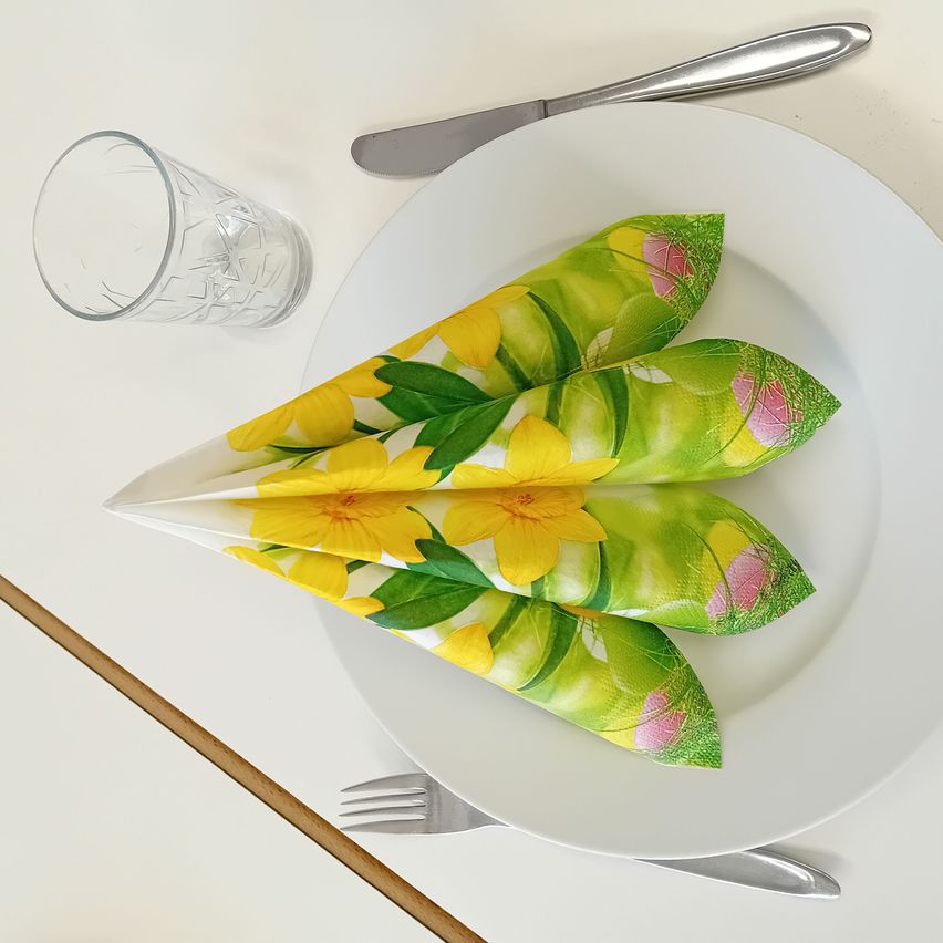 Ein weißer Teller mit zwei gefalteten gelben und grünen Blumenservietten. Ein Messer, eine Gabel und ein Glas sind in der Nähe. Die Tischdekoration ist auf einem weißen Tisch.