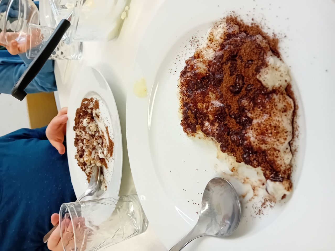 Zwei Teller mit Dessert stehen auf einem Tisch. Auf einem Teller sind eine Gabel und ein Glas in der Nähe. Das Dessert ist mit Schokolade überzogen und mit gepulverter Schokolade bestreut. Der andere Teller hat weniger Dessert.
