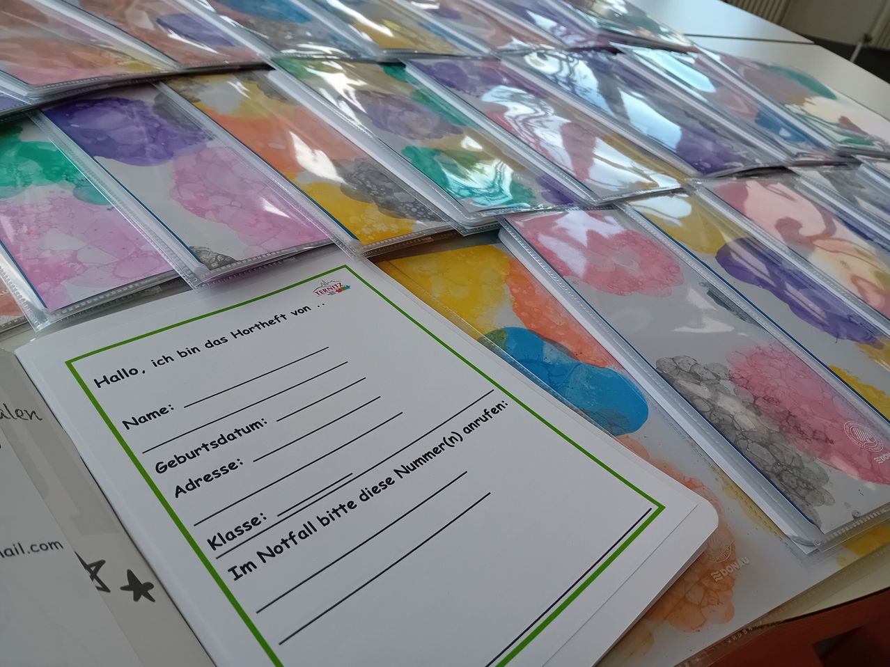 A table with several transparent plastic folders, each containing a colorful, abstract watercolor painting. A white form with green borders is partially visible, featuring text in German asking for personal information.