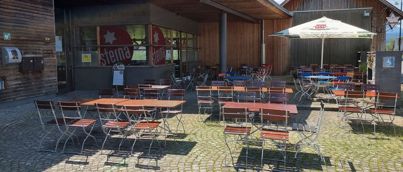 Eine Außensitzfläche eines Restaurants mit Holztischen und Stühlen, einem Schild mit der Aufschrift 'Stena' und einem großen Schirm. Das Gebäude hat eine Holzfassade und Glasfenster.