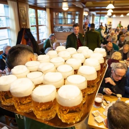 Ein überfüllter Biergarten mit einem Kellner, der ein Tablett mit Bierkrügen trägt, während Gäste an Tischen sitzen und plaudern.