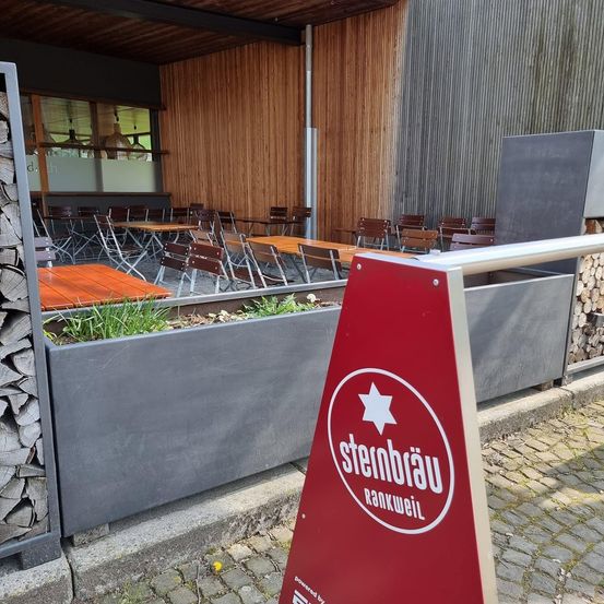 Ein rotes dreieckiges Schild mit dem Text 'Stembrau' und einem weißen Stern in der Mitte steht auf einem gepflasterten Weg. Dahinter ist ein Restaurant mit Holzwänden und Tischen und Stühlen zu sehen.