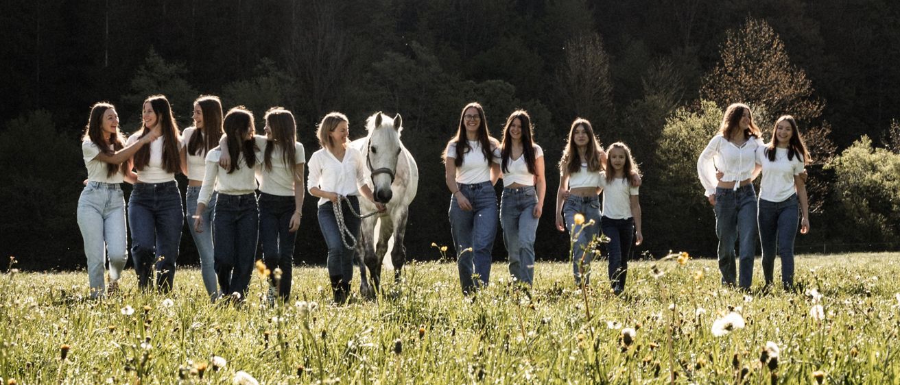 Eine Gruppe von Frauen geht mit einem weißen Pferd in einem Feld unter einem hellen Sonnenschein, umgeben von grünem Gras und Bäumen.