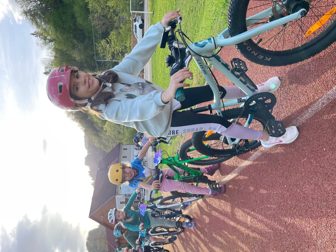 Ein Mädchen mit einem pinken Helm fährt mit ihren Freunden auf einer roten Strecke ein Fahrrad.