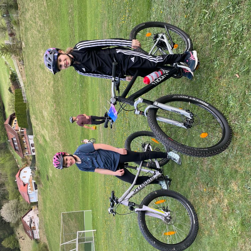 Zwei Kinder posieren mit ihren Fahrrädern auf einer Grasfläche und tragen Helme. Ein Kind steht, während das andere sitzt. Im Hintergrund befinden sich Häuser und ein Zaun.