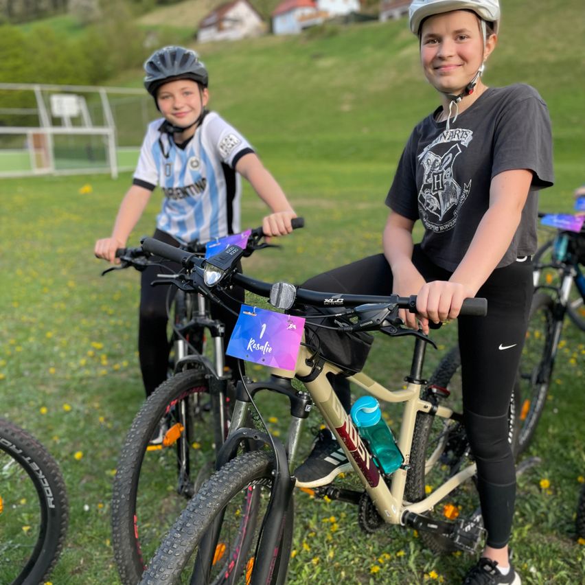Zwei junge Radfahrer in Helmen und Radsporttrikots posieren für ein Foto auf einem Rasenfeld. Der Junge links trägt ein Argentinien-Trikot, während das Mädchen rechts ein schwarzes Shirt mit einem Hogwarts-Emblem trägt.