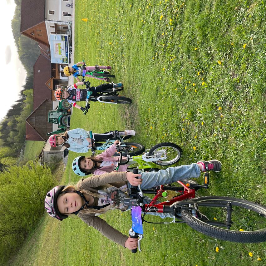 Fünf Kinder mit Helmen fahren auf einem Rasenfeld mit Fahrrädern, mit Gebäuden und Bäumen im Hintergrund.