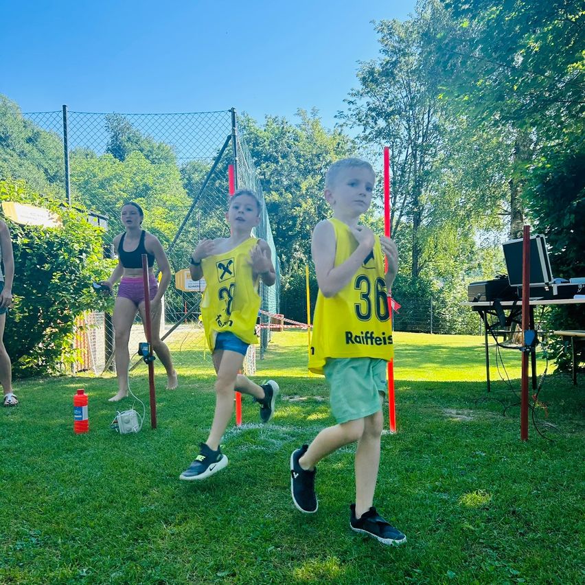 Drei Kinder laufen mit gelben Westen über ein Grasfeld. Zwei sind mit 36 und 37 nummeriert. Eine Frau in einem schwarzen Bikini ist im Hintergrund.