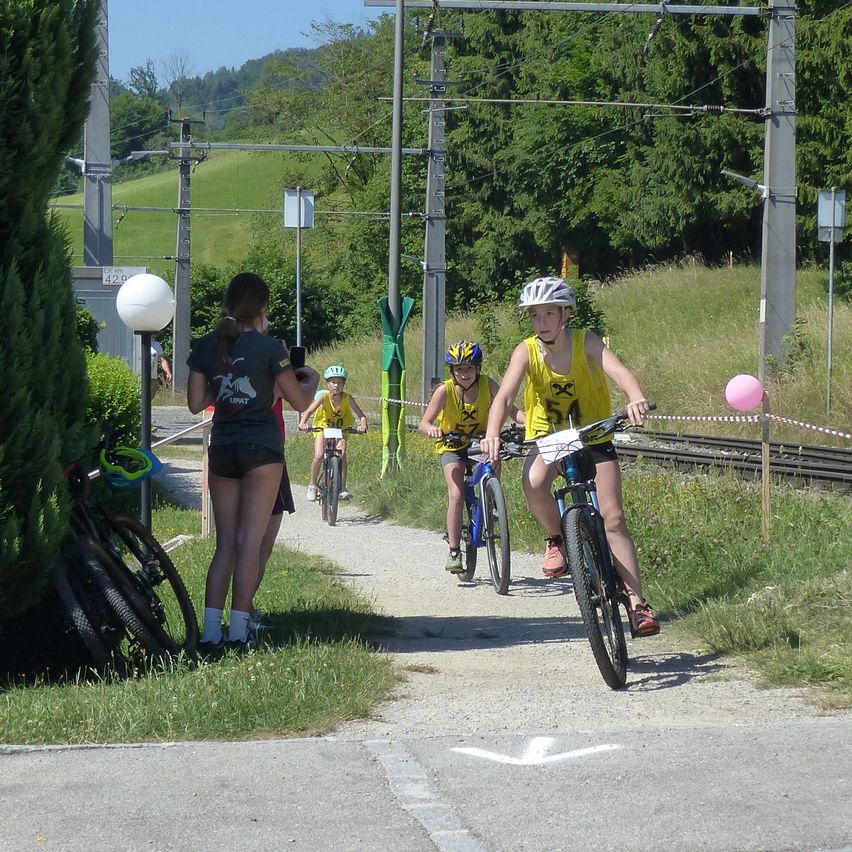 Drei Radfahrer mit Helmen fahren auf einem Pfad in der Nähe einer Eisenbahnstrecke. Eine Frau steht in der Nähe, und zwei Fahrräder sind auf dem Gras geparkt.