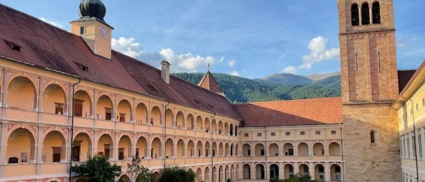 Ein Klosterhof mit einem großen, gewölbten Gebäude und einem hohen Glockenturm. Der Hof ist von einem üppigen Rasen mit Pflanzen und Bäumen umgeben. Das Kloster ist aus Stein gebaut und hat einen Glockenturm. Im Hintergrund befinden sich Berge.