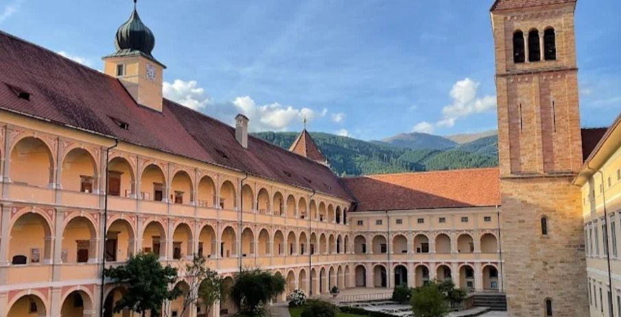 Ein Klosterhof mit einem großen, gewölbten Gebäude und einem hohen Glockenturm. Der Hof ist von einem üppigen Rasen mit Pflanzen und Bäumen umgeben. Das Kloster ist aus Stein gebaut und hat einen Glockenturm. Im Hintergrund befinden sich Berge.