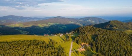 Luftaufnahme eines Berggebiets mit einer gewundenen Straße und verstreuten Häusern unter einem blauen Himmel mit einigen Wolken.