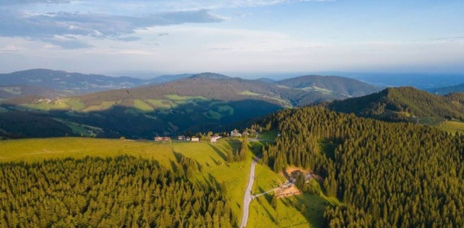 Luftaufnahme eines Berggebiets mit einer gewundenen Straße und verstreuten Häusern unter einem blauen Himmel mit einigen Wolken.