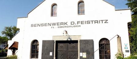 Ein weißes Gebäude mit Rundbogenfenstern und einer großen Holztür. Es steht 'Sensenwerk D.Feistritz FL.Obergruber' an der Wand. In der Nähe der Tür befindet sich ein kleines Schild.