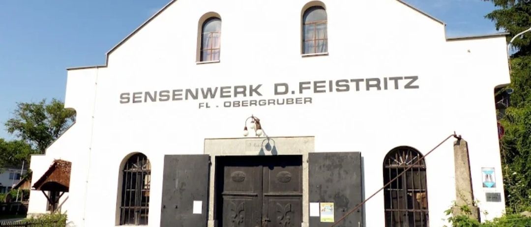 Ein weißes Gebäude mit Rundbogenfenstern und einer großen Holztür. Es steht 'Sensenwerk D.Feistritz FL.Obergruber' an der Wand. In der Nähe der Tür befindet sich ein kleines Schild.