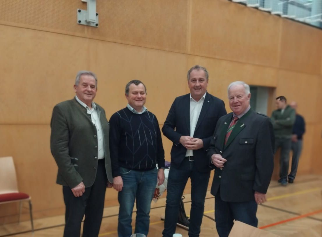 Vier Männer stehen lächelnd in einer Turnhalle. Sie tragen formelle und lässige Kleidung. Hinter ihnen befindet sich eine Holzwand mit einer weißen Sicherheitskamera.