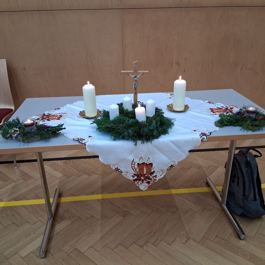 Ein Tisch mit weißer Tischdecke und einem Kreuz, geschmückt mit Kerzen und Girlanden, steht in einem Raum. Ein Stuhl und ein Rucksack sind in der Nähe.