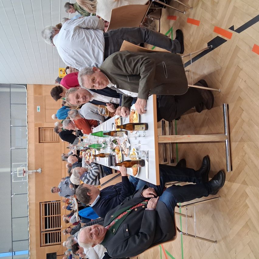 Eine Gruppe von Menschen sitzt an einem langen Tisch mit verschiedenen Speisen und Getränken darauf. Sie sind in einem Saal mit Holzböden und einige von ihnen tragen Krawatten. Der Raum hat einen Basketballkorb und eine Tür.