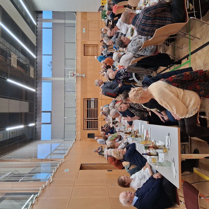 Eine Indoor-Versammlung mit mehreren Personen, die um Tische herum sitzen, möglicherweise für eine Mahlzeit oder Veranstaltung, in einer Halle mit Basketballkorb und Glasfenstern.