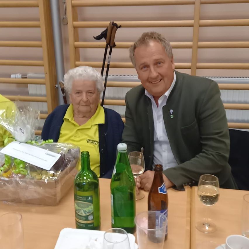 Ein Mann und eine ältere Frau sitzen an einem Tisch mit Weingläsern, einer Flasche und einem Korb mit Obst. Sie lächeln und scheinen sich bei einer Veranstaltung zu befinden. Dahinter ist eine Holzwand und ein Heizkörper zu sehen.