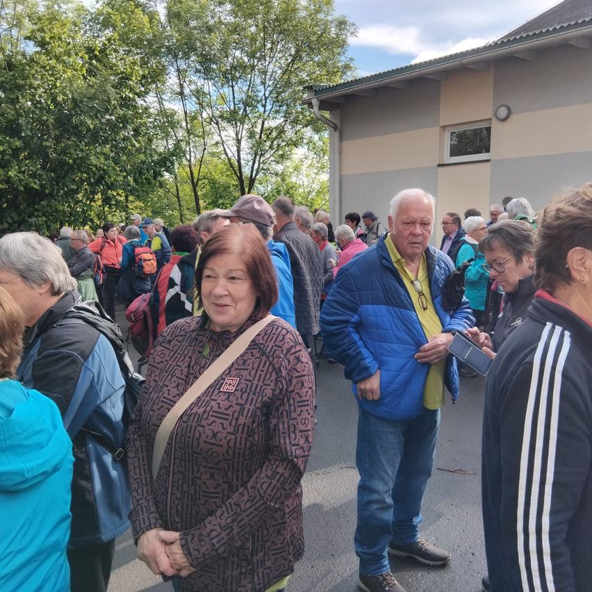 Eine Gruppe von Menschen, darunter eine Frau in einer braunen Jacke, steht vor einem Gebäude. Einige tragen Brillen und Rucksäcke.