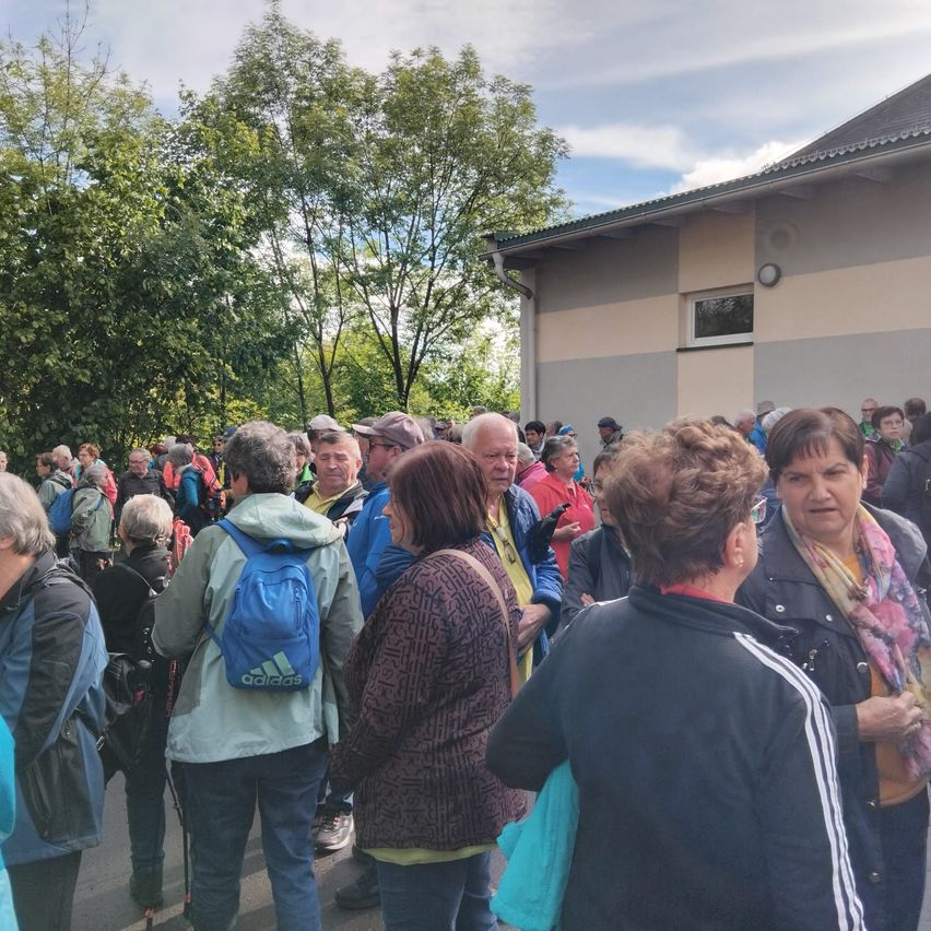 Eine Gruppe älterer Menschen versammelt sich vor einem Gebäude, einige tragen Rucksäcke. Sie sind in ein Gespräch vertieft. Bäume und ein bewölkter Himmel sind im Hintergrund zu sehen.