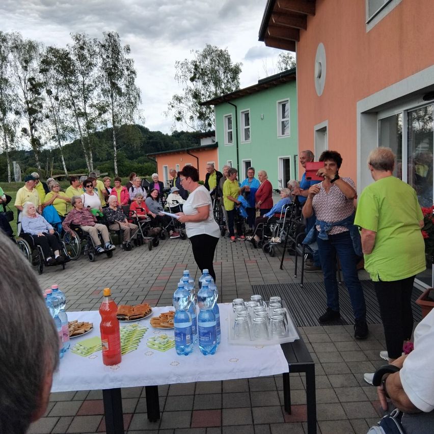 A group of elderly people in wheelchairs gathered outside a building with several people taking pictures. A table with refreshments and water bottles is set up.