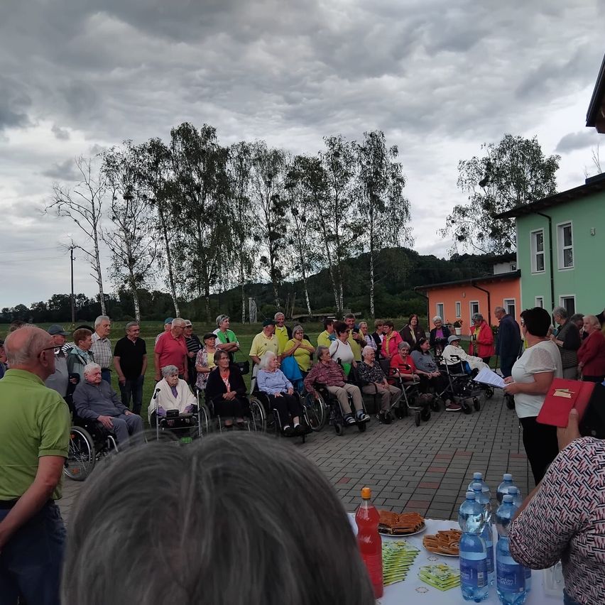 A group of elderly people, some in wheelchairs, gathers outside under a cloudy sky. Nearby, there is a table with bottles and food. Trees and buildings are visible in the background.