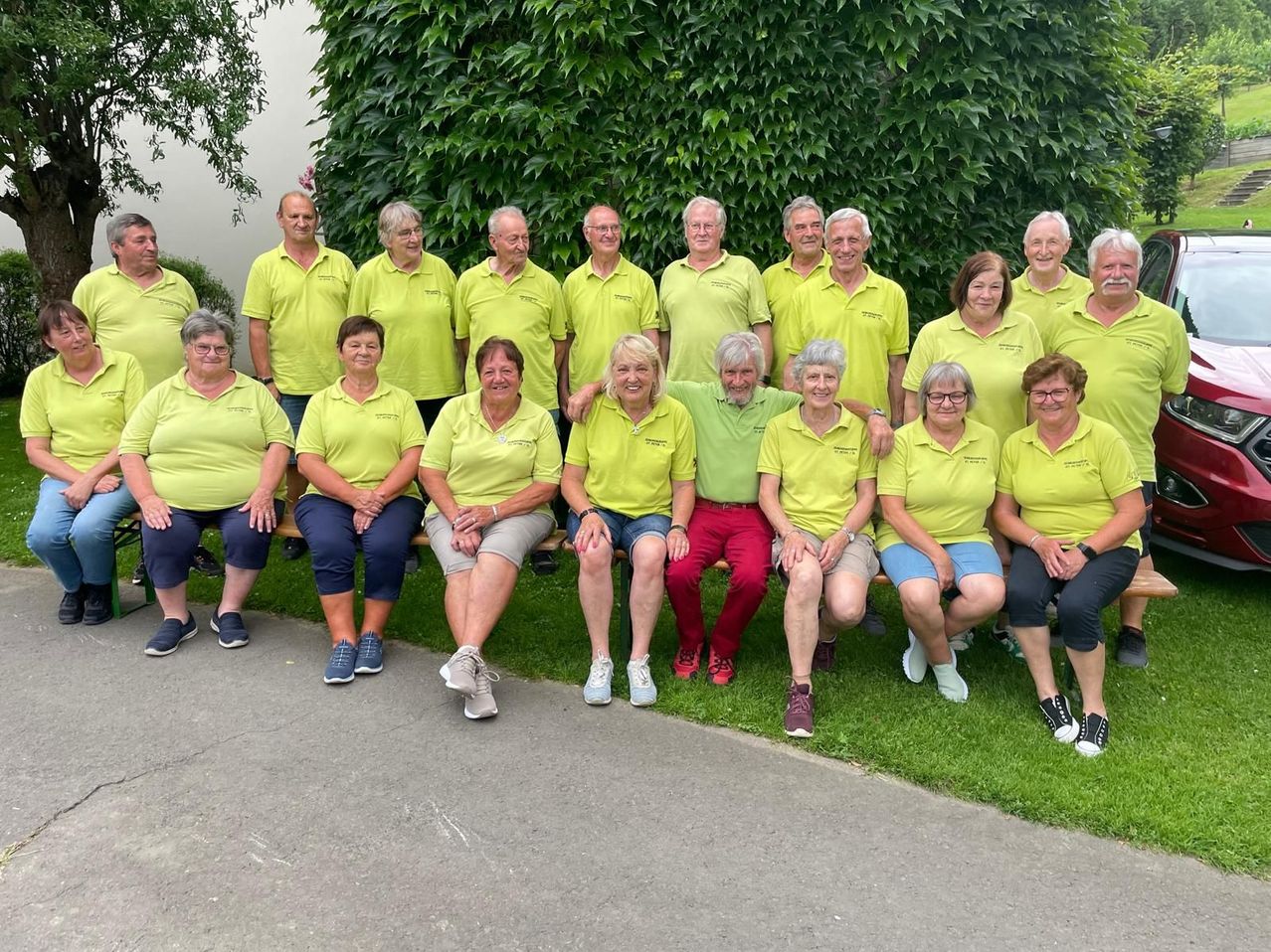 Eine Gruppe von Erwachsenen in gelben Shirts und roten Hosen posiert für ein Foto im Park. Sie sitzen und stehen auf dem Rasen.