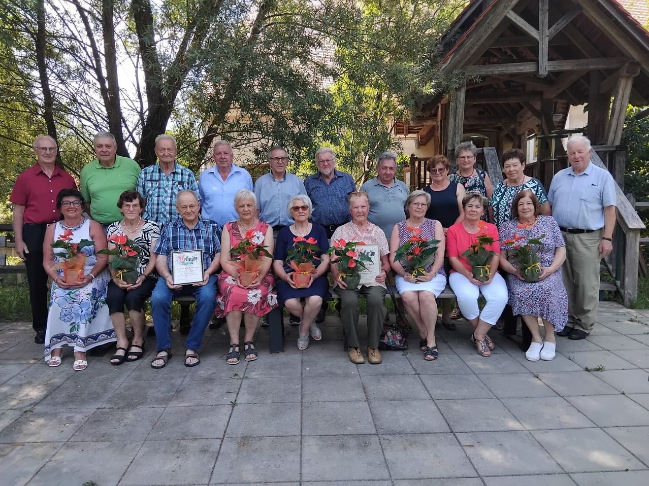 Eine Gruppe von Menschen, einige mit Blumen, sitzt auf einer Bank unter einem Pavillon. Sie sind lässig gekleidet. Einige tragen Brillen und Halsketten. Einige von ihnen halten ein Zertifikat.