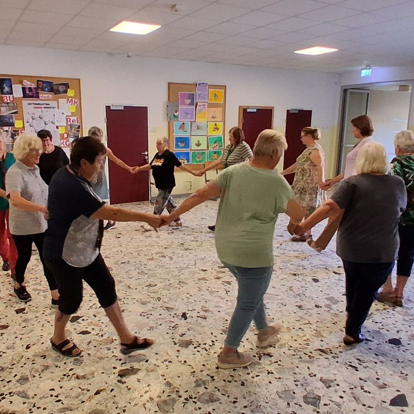 A group of elderly women are holding hands and dancing in a circle in a room with white walls and a speckled floor.