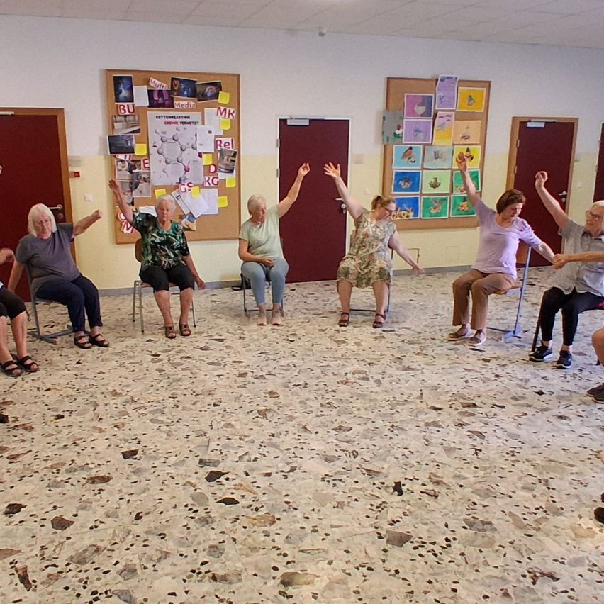 A group of elderly women are sitting in chairs and raising their hands in a room with bulletin boards and doors.