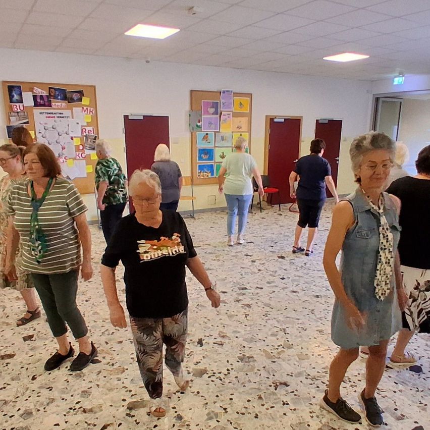 A group of elderly women are walking in a room with a marble floor and bulletin boards on the wall. Some are wearing glasses and sneakers.