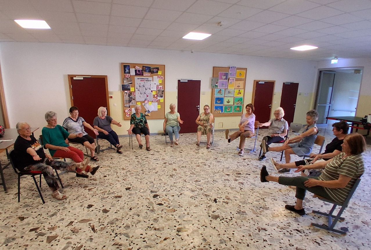 A group of elderly women are sitting in a circle, possibly in a community center, wearing casual clothing. They are engaged in an activity, possibly a meeting or a discussion. The room has a white wall with bulletin boards and a ceiling with lights.