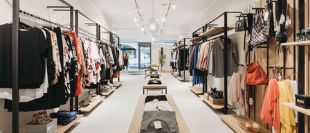 Ein sauberer, gut beleuchteter Bekleidungsladen mit Kleidern an Kleiderbügeln und ausgestellten Schuhen. Der Laden hat ein modernes, minimalistisches Design mit weißen Wänden und Holzregalen.