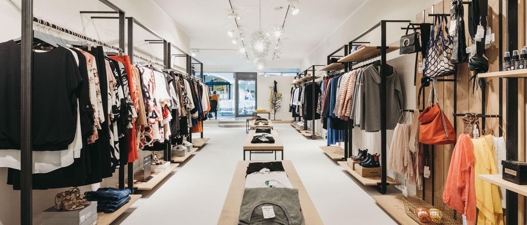 Ein sauberer, gut beleuchteter Bekleidungsladen mit Kleidern an Kleiderbügeln und ausgestellten Schuhen. Der Laden hat ein modernes, minimalistisches Design mit weißen Wänden und Holzregalen.