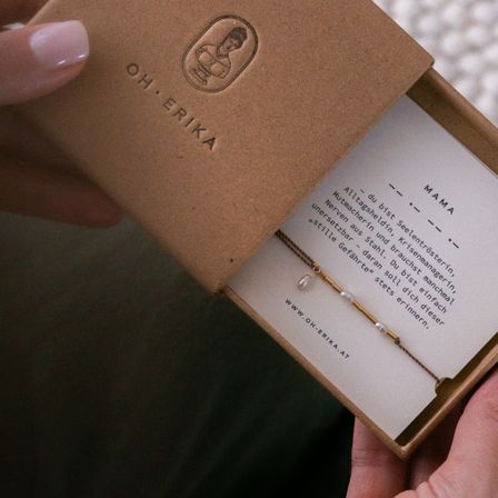 Eine Hand öffnet eine braune Schachtel mit 'Oh Erika', darin befindet sich eine Perlenkette mit einer Karte, die 'Mama' besagt.