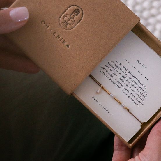 Eine Hand öffnet eine braune Schachtel mit 'Oh Erika', darin befindet sich eine Perlenkette mit einer Karte, die 'Mama' besagt.