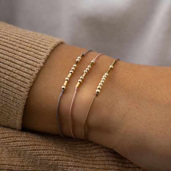 Eine Person mit einem braunen Pullover und drei goldenen Perlenarmbändern am Handgelenk.