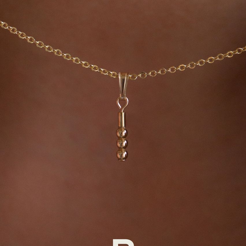 Eine goldene Kette mit einem Anhänger, der drei kleine goldene Perlen aufweist, die an einem Haken hängen. Die Kette ist vor einem braunen Hintergrund platziert.
