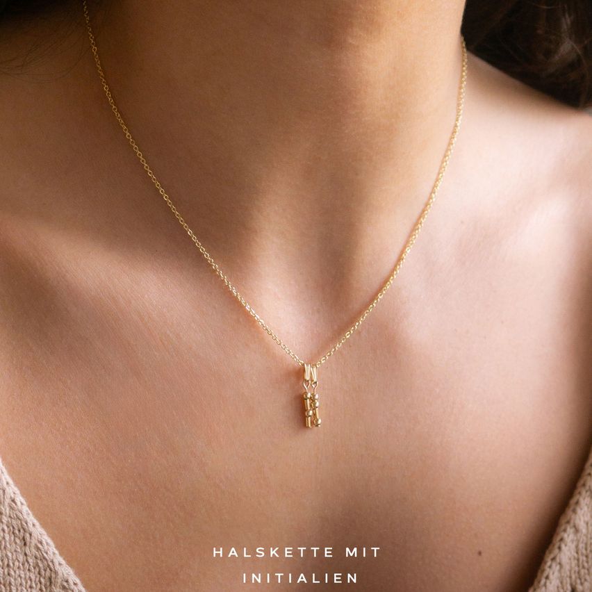 Eine Frau trägt eine goldene Halskette mit kleinem Anhänger auf ihrem Hals. Unten steht 'Halskette mit Initialen'.